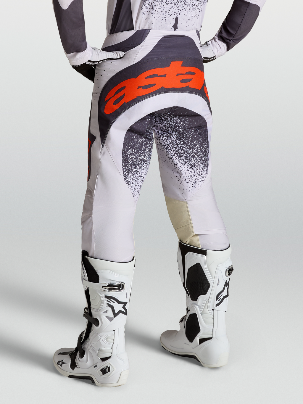 Alpinestars Techstar Swan, pantalon de motocross pour homme, lilas clair et gris foncé avec accents rouge fluo, porté avec des bottes Tech 10 blanches, idéal pour le racing off-road