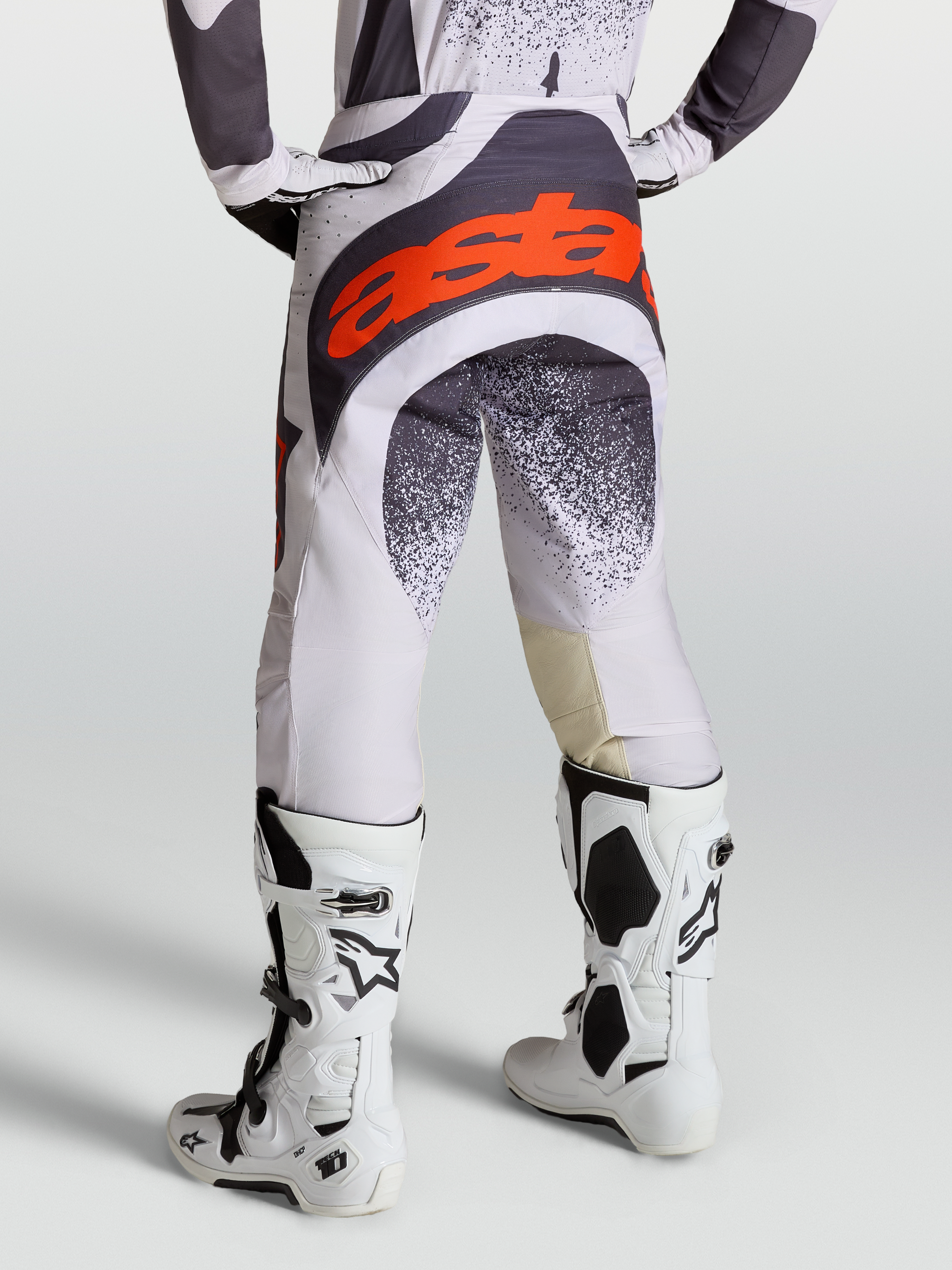 Alpinestars Techstar Swan, pantalon de motocross pour homme, lilas clair et gris foncé avec accents rouge fluo, porté avec des bottes Tech 10 blanches, idéal pour le racing off-road