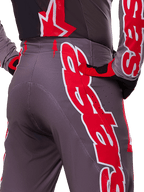 Alpinestars 2025 Supertech Lipan Pants, pantalon de motocross, Smoke Bright Red, vue rapprochée de l'arrière montrant un tissu gris foncé avec des graphismes rouges vifs, porté avec des gants et un maillot rouges et noirs assortis, doté de perforations de ventilation et de coutures ergonomiques pour des performances tout-terrain