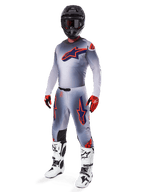 Pilote de motocross portant la tenue de course complète Alpinestars Supertech Lipan 2025, gris et orange fluo, maillot gris avec grand logo orange sur la poitrine, pantalon assorti avec accents orange, casque noir, gants rouges et bottes blanches, équipement de course tout-terrain professionnel