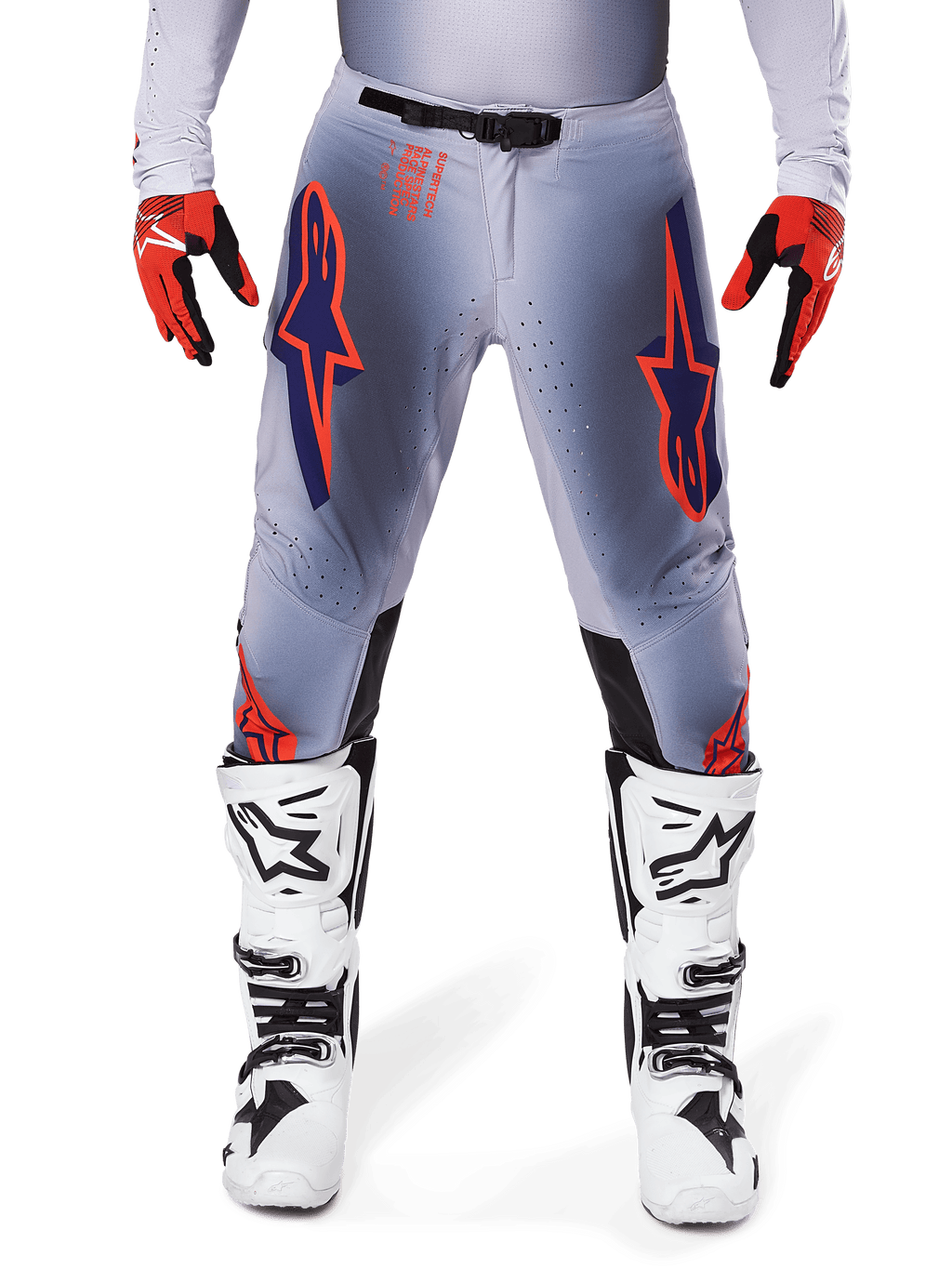 Alpinestars 2025 Supertech Lipan Pants, pantalon de motocross, gris et orange fluo, porté avec des gants rouges et noirs et des bottes blanches et noires, arborant de grands logos étoilés Alpinestars sur les cuisses et des perforations de ventilation pour la compétition de niveau professionnel.