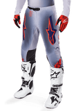 Pantalon Alpinestars Supertech Lipan 2025, pantalon de motocross, gris et orange fluo, avec motifs d'étoiles bleu marine et empiècements noirs à l'intérieur des genoux, porté avec des gants rouge-orange et des bottes blanches, équipement de course tout-terrain professionnel