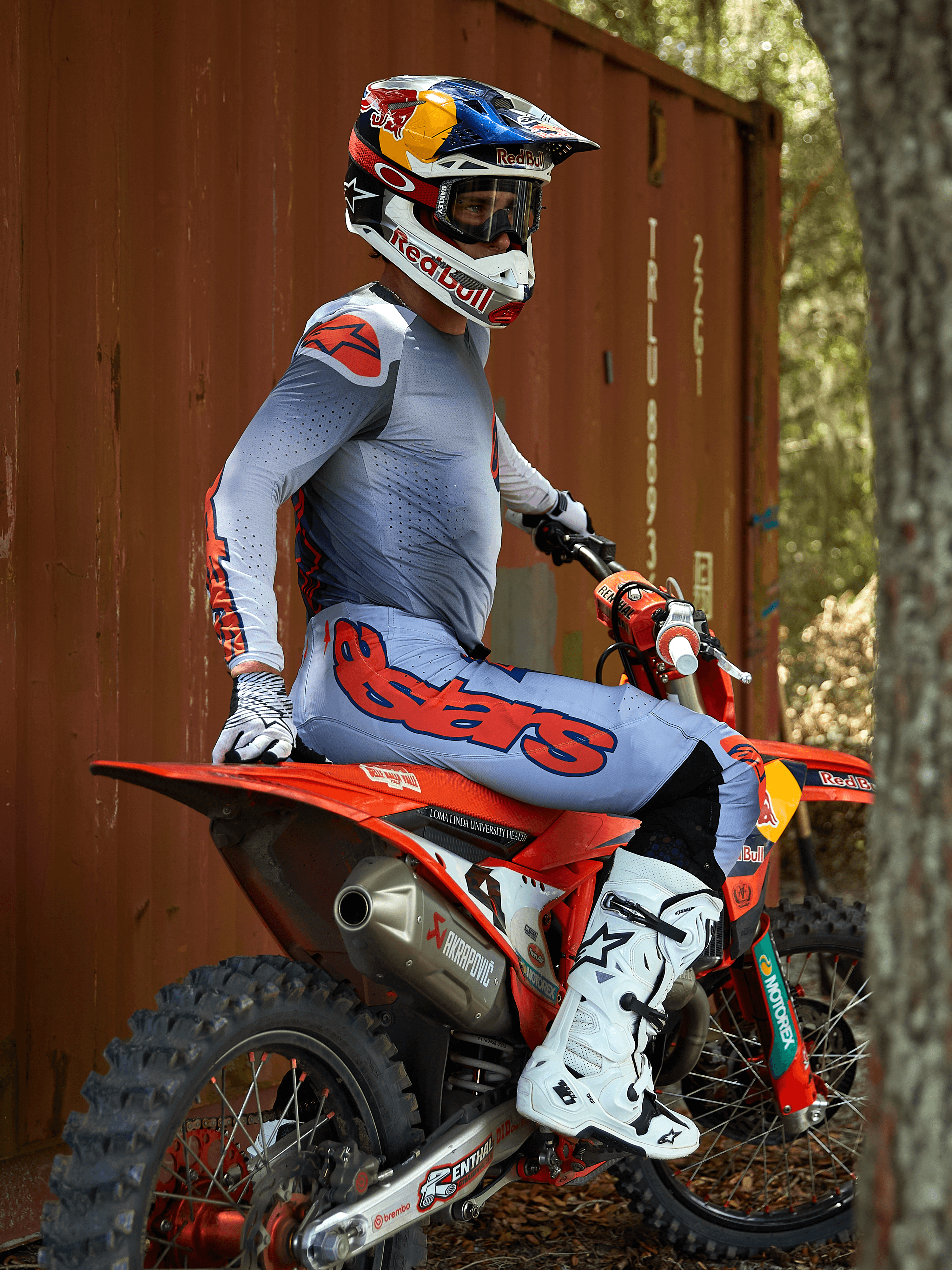 Pilote de motocross assis sur une moto tout-terrain rouge, portant l'ensemble d'équipement Alpinestars Supertech Lipan 2025 en gris et orange fluo, pantalon bleu clair avec logo Astars orange, maillot assorti, bottes de motocross blanches et casque Red Bull, équipement de course professionnel coordonné