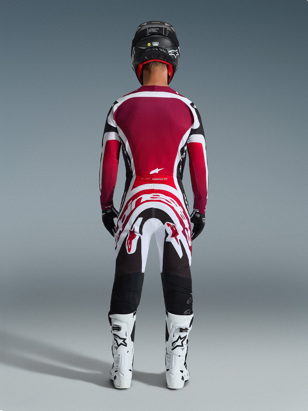 Motocross rider from behind wearing Alpinestars 2026 Supertech AFD Vista Pants, Black Red White, paired with a matching jersey, white racing boots, and black helmet, complete professional gear set for off-road racing
