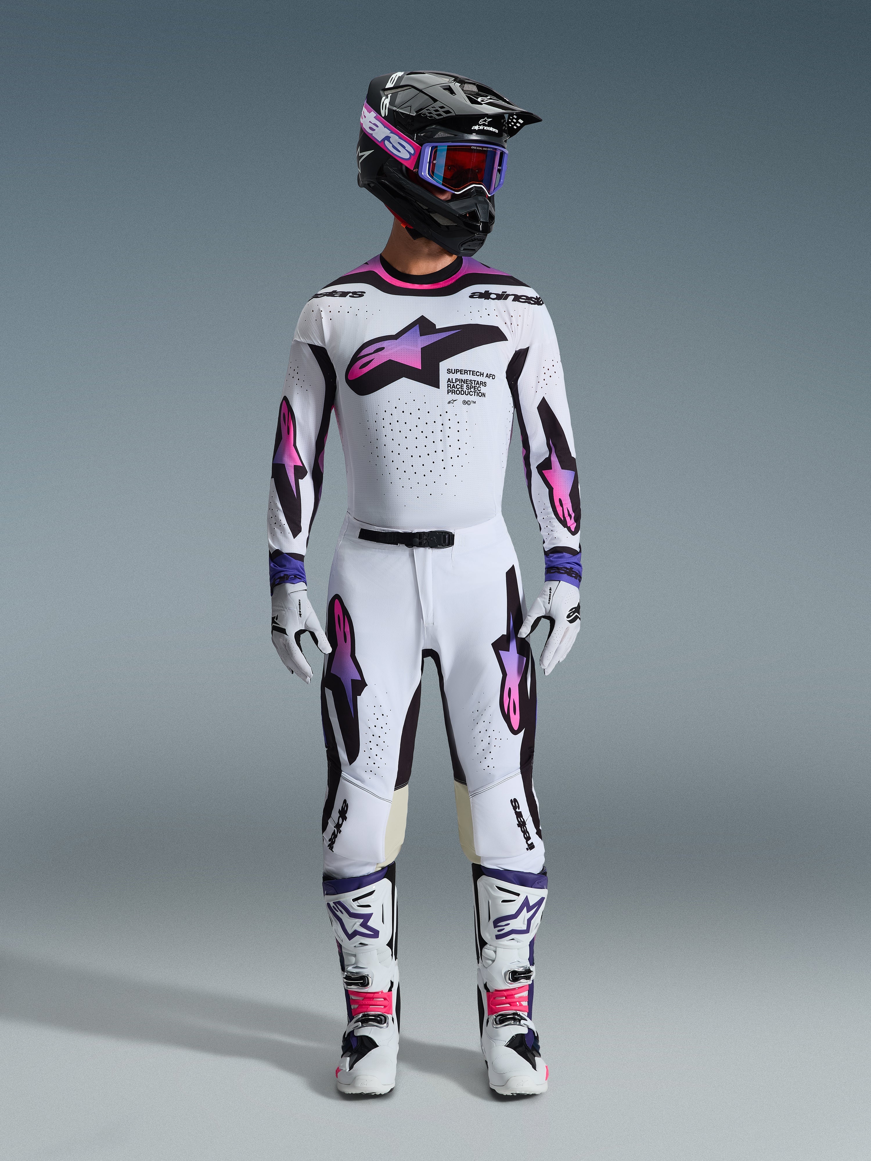 Motocross rider wearing the Alpinestars 2026 Supertech AFD Vista Gear Set, white jersey and pants with pink and purple gradient star graphics, black helmet with matching goggles, white boots with pink accents, and white gloves, coordinated professional off-road racing outfit