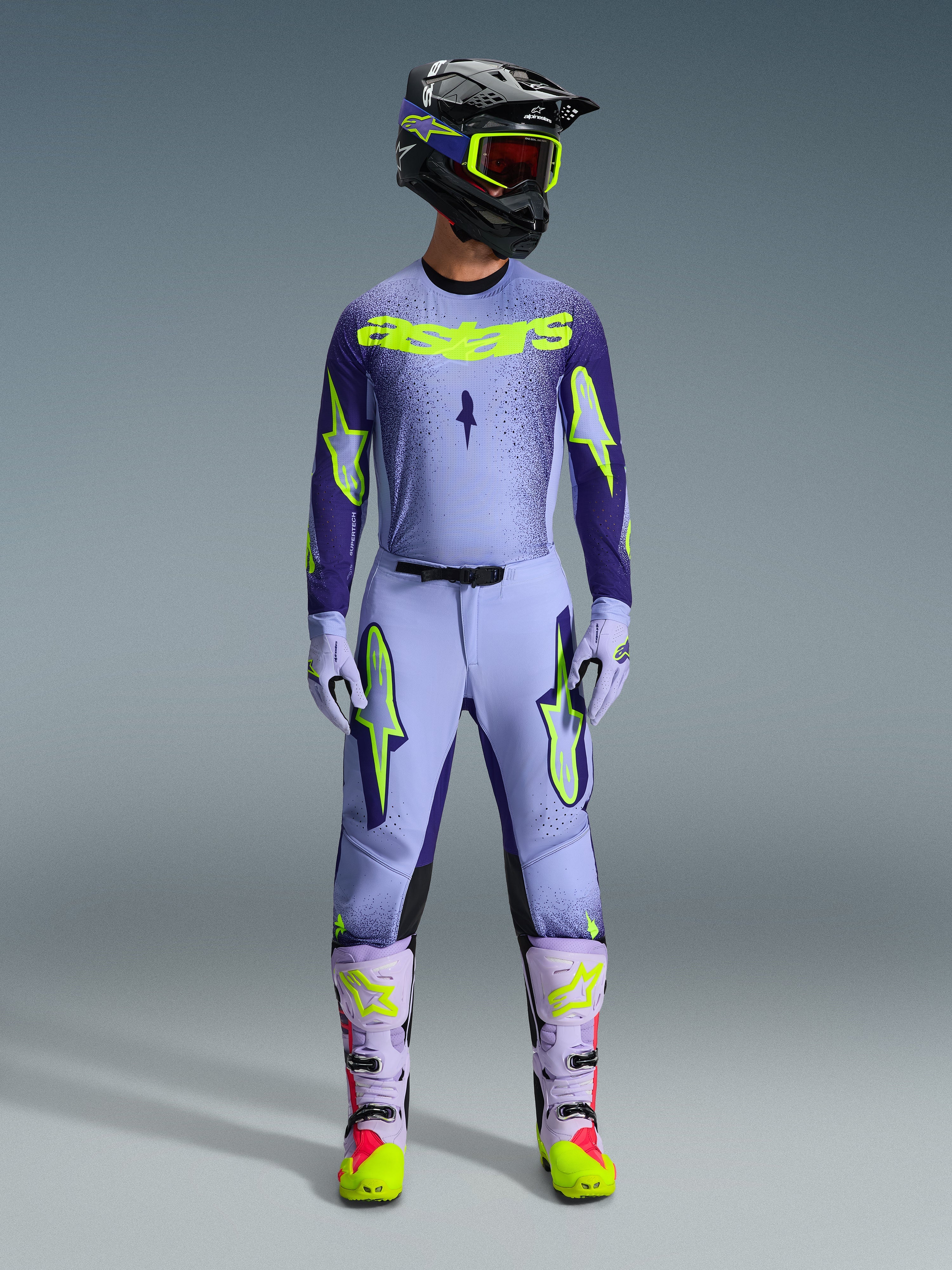 Ensemble d'équipement Alpinestars 2026 Supertech Scenz, tenue de course de motocross, violet clair et jaune-vert néon, équipement de protection complet comprenant un casque noir avec des accents violets, un maillot avec un marquage jaune néon audacieux, un pantalon assorti avec des graphismes d'étoiles et des bottes coordonnées avec des touches roses.