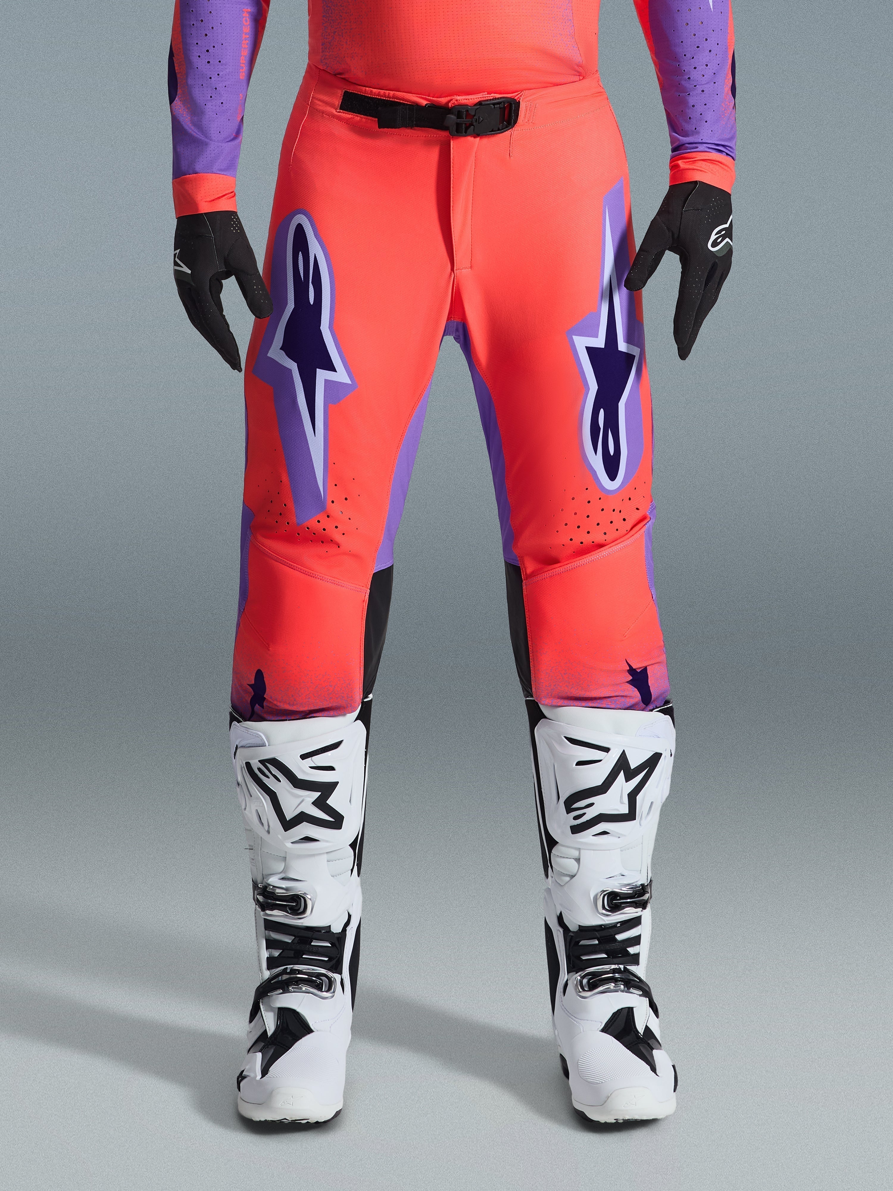 Alpinestars 2026 Supertech Scenz Pants, pantalon de motocross, corail vif et violet, porté avec un maillot assorti, des bottes blanches et noires et des gants noirs, avec de grands logos Astars violets sur les cuisses et une boucle de taille magnétique noire, coupe athlétique professionnelle pour la course