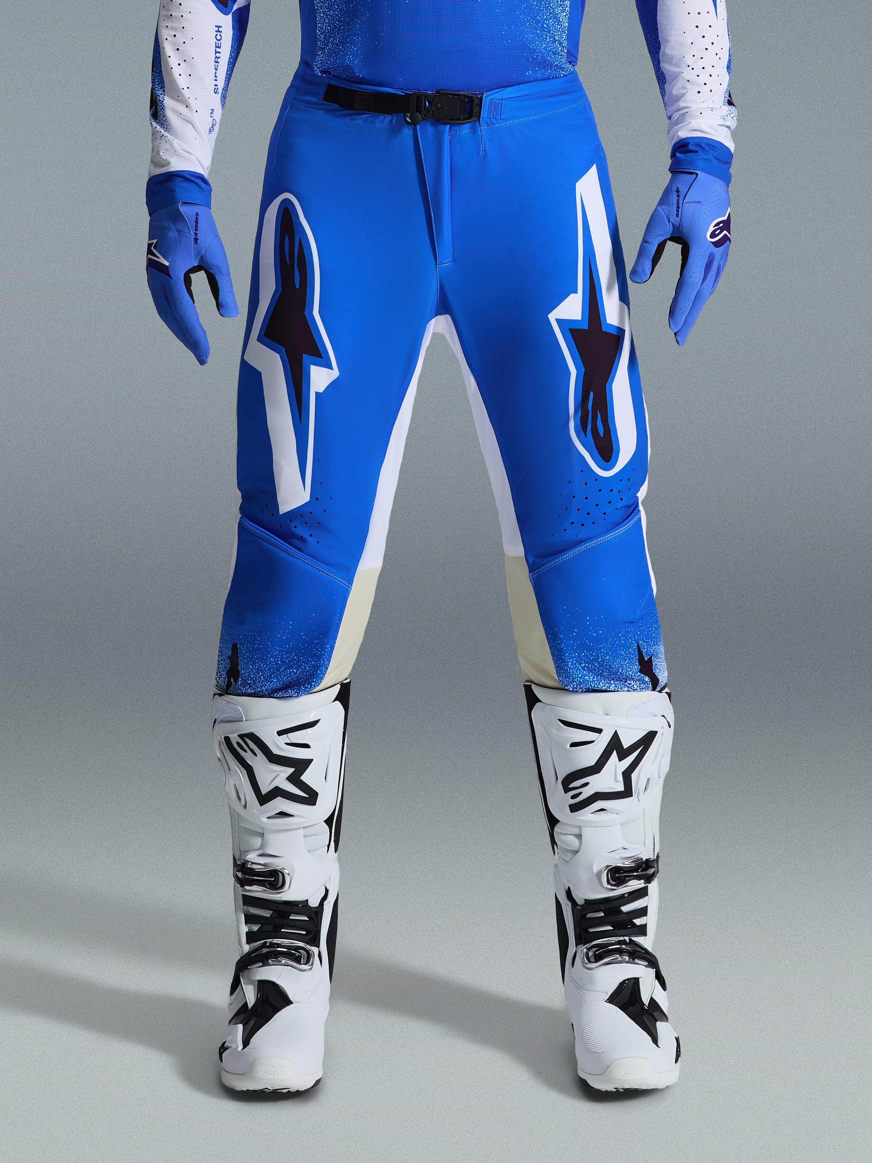 Pantalon Alpinestars Supertech Scenz 2026, pantalon de motocross, bleu UCLA et blanc, porté avec des gants bleus assortis et des bottes blanches, avec de grands graphismes blancs et noirs sur les cuisses, coupe athlétique pour une performance tout-terrain