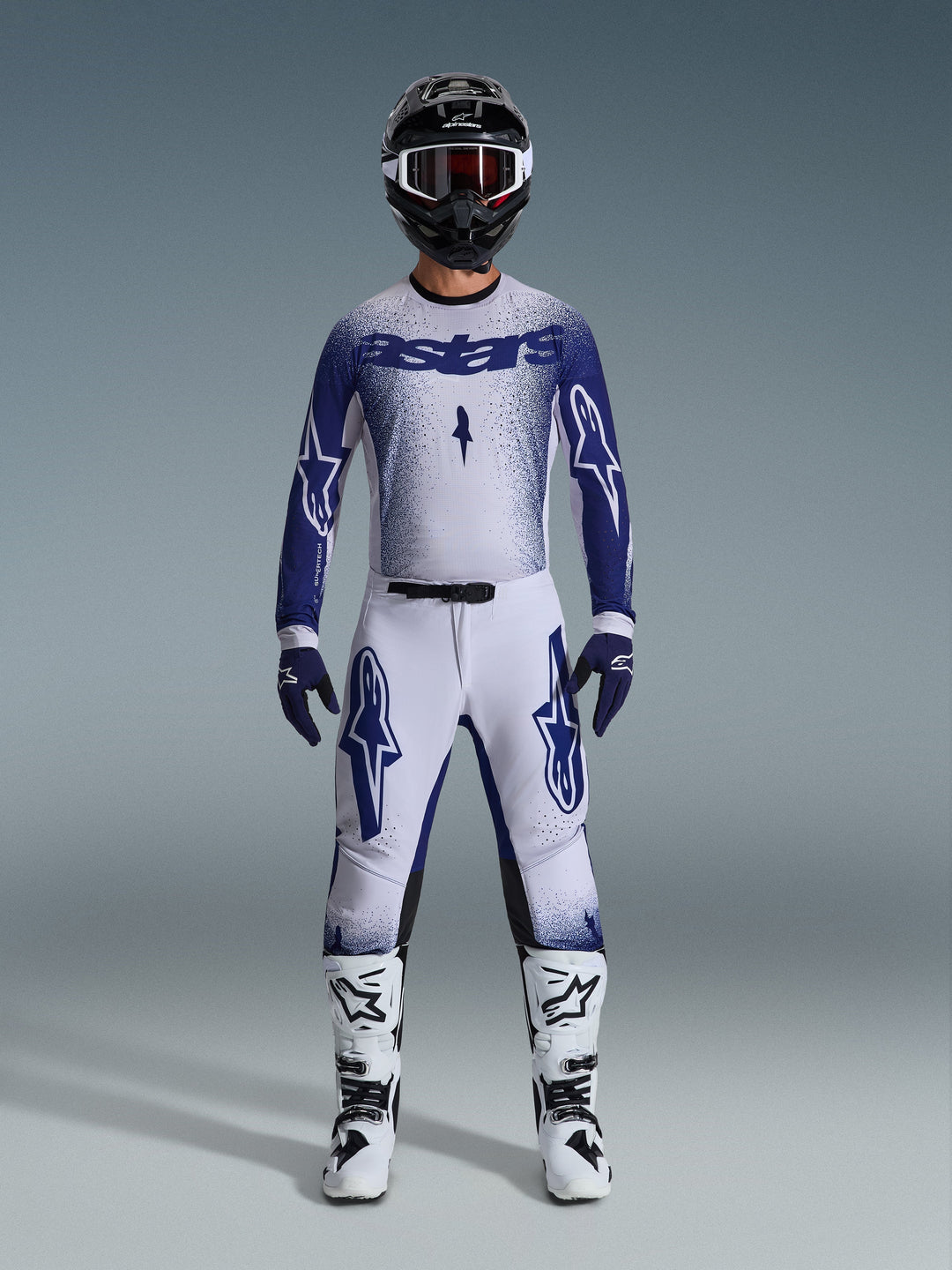 Pilote de motocross portant l'ensemble Alpinestars Supertech Scenz 2026, maillot blanc et bleu foncé avec pantalon assorti, orné de grands logos Astars et d'un design graphique moucheté, avec un casque noir, des bottes de protection blanches et noires et des gants bleus coordonnés, tenue de course professionnelle complète