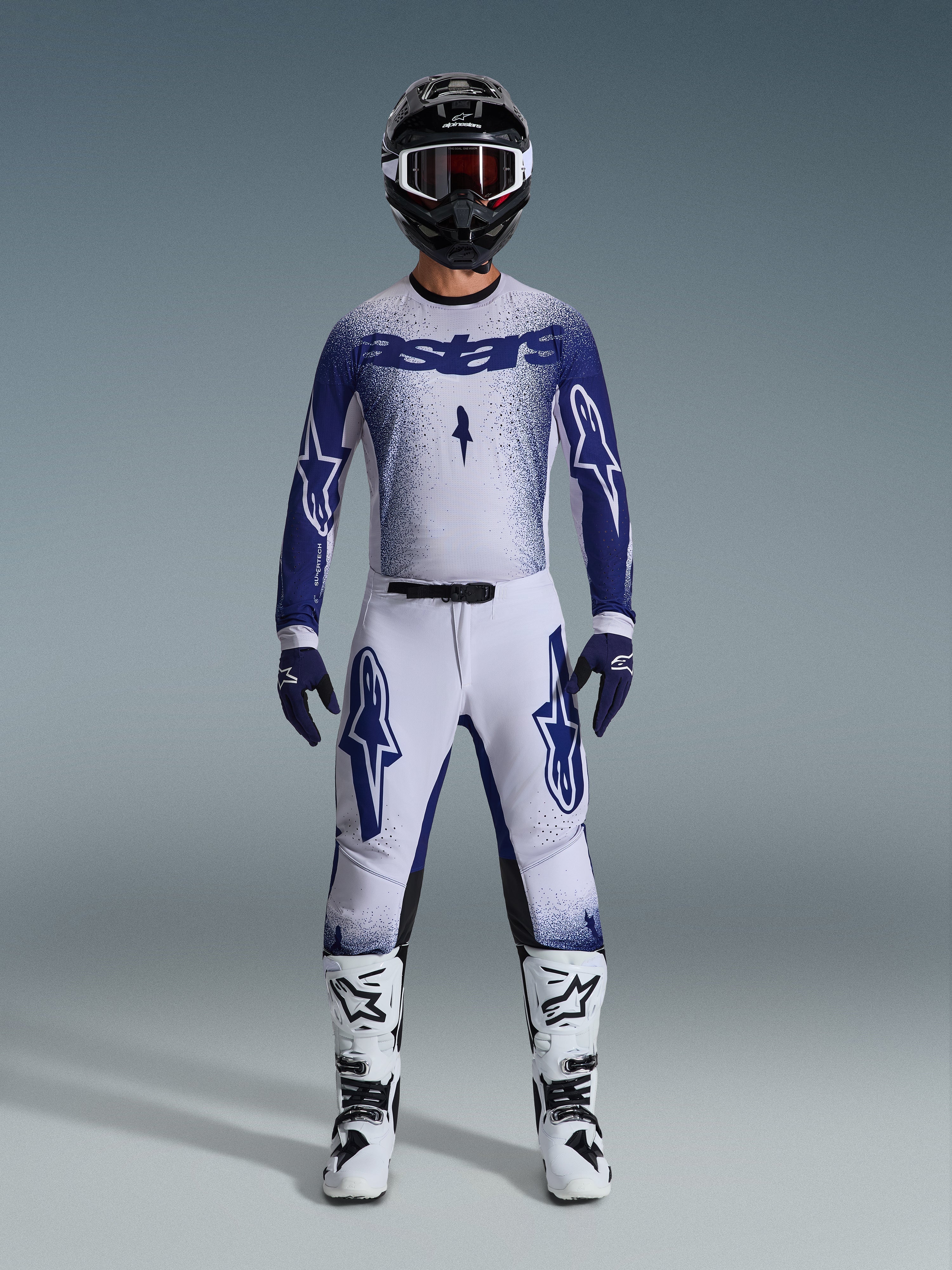 Pilote de motocross portant l'ensemble Alpinestars Supertech Scenz 2026, maillot blanc et bleu foncé avec pantalon assorti, orné de grands logos Astars et d'un design graphique moucheté, avec un casque noir, des bottes de protection blanches et noires et des gants bleus coordonnés, tenue de course professionnelle complète