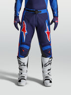 Alpinestars Pantalon Racer Hunt, pantalon de motocross pour homme, bleu et rouge, construction pré-courbée avec graphismes étoilés, porté avec des bottes blanches