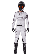Pilote de motocross portant le pantalon Alpinestars 2025 Racer Hollow en Light Camo Brown, associé à un maillot à manches longues assorti blanc et gris, un casque noir avec lunettes orange-rouge, des gants noirs et des bottes noires, ensemble de course coordonné pour la performance tout-terrain.