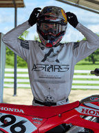 Pilote de motocross portant l'équipement Alpinestars 2025 Racer Hollow, maillot camouflage clair avec marquage ASTARS imposant, ajustant un casque Red Bull muni de lunettes transparentes, debout à côté d'une moto Honda rouge dans un cadre extérieur tout-terrain
