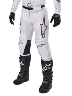 Alpinestars 2025 Racer Hollow Pants, pantalon de motocross, camouflage blanc et gris, avec de grands logos étoiles aux contours noirs et des empiècements en cuir noir à l'intérieur des genoux, porté avec des bottes et des gants de motocross noirs, conception durable pour la course tout-terrain.