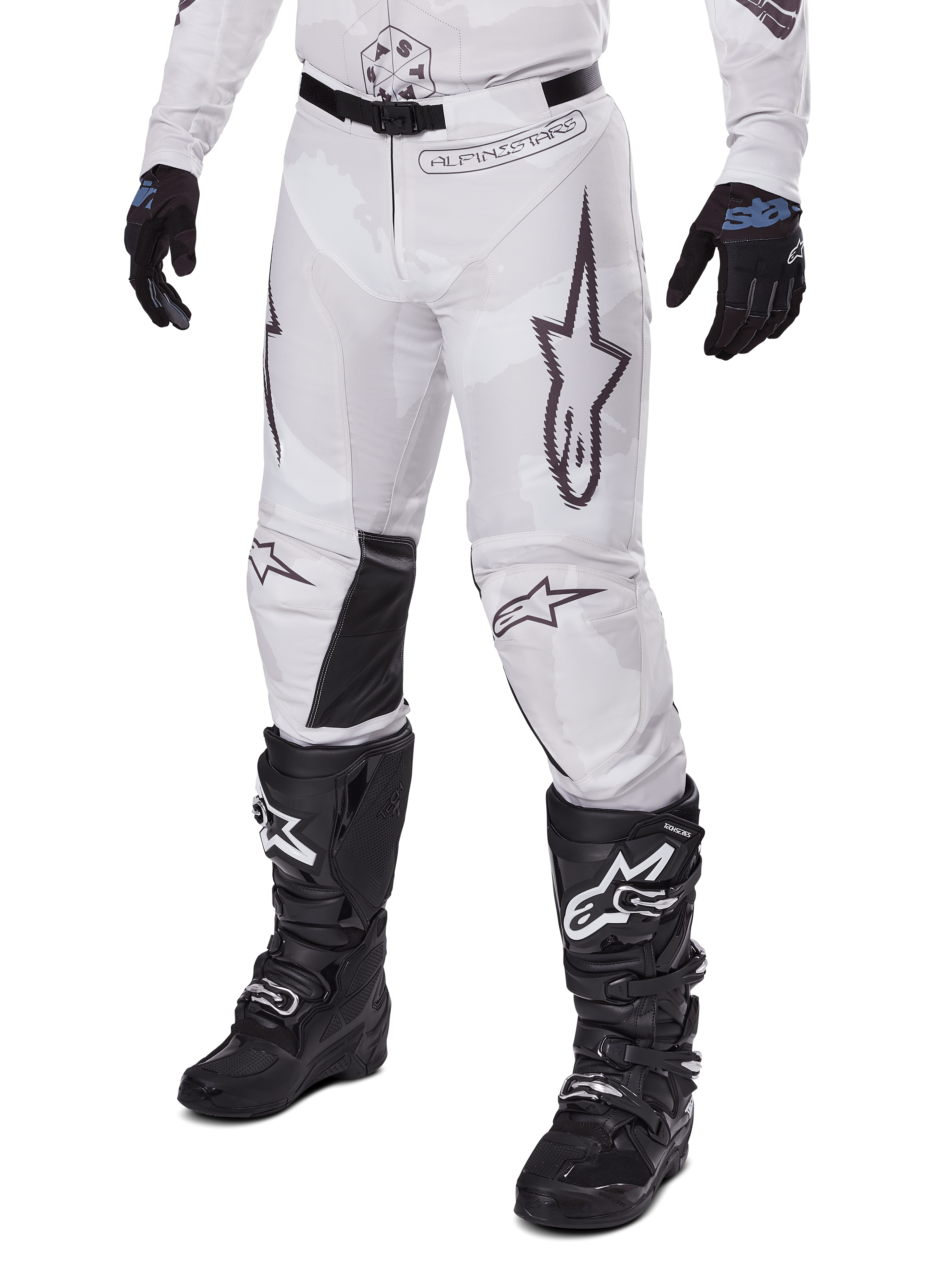 Alpinestars 2025 Racer Hollow Pants, pantalon de motocross, camouflage blanc et gris, avec de grands logos étoiles aux contours noirs et des empiècements en cuir noir à l'intérieur des genoux, porté avec des bottes et des gants de motocross noirs, conception durable pour la course tout-terrain.