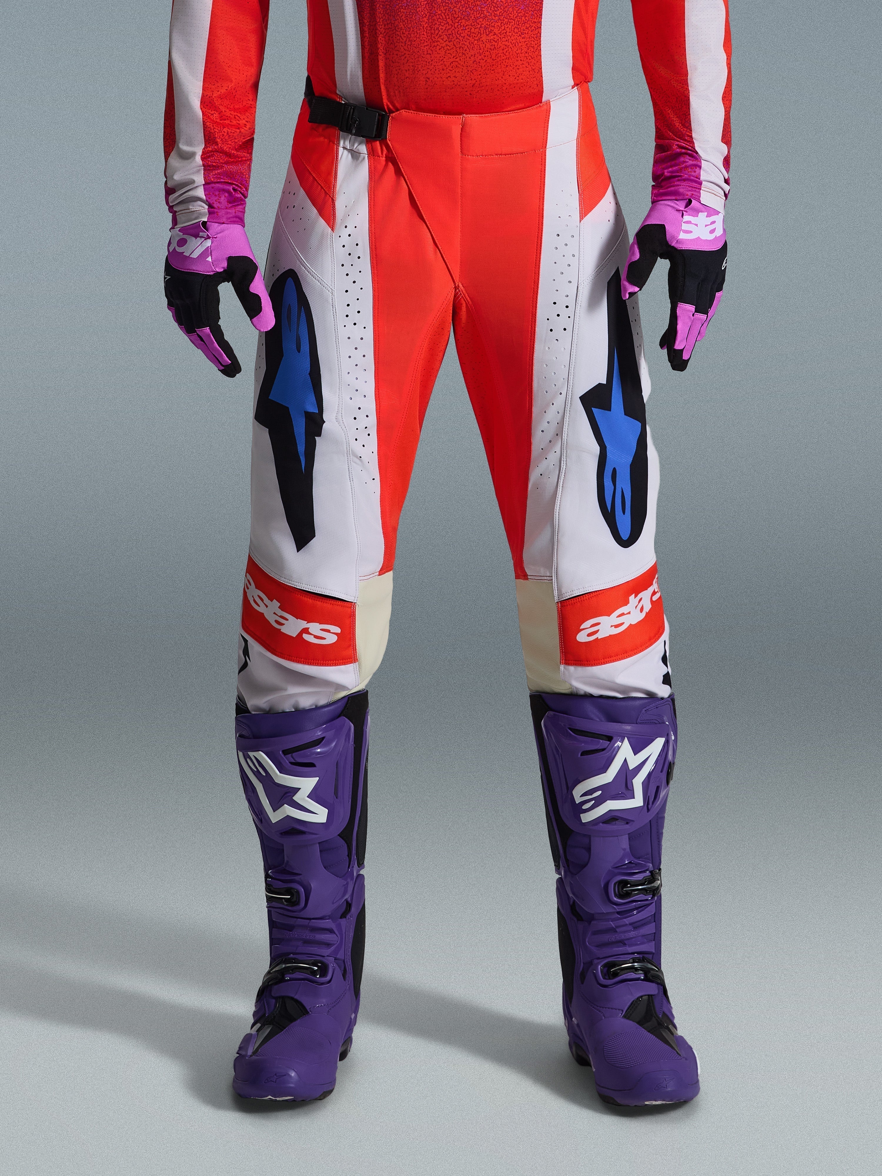 Alpinestars 2026 Techstar Knif Pants, pantalon de motocross orange fluo et gris clair avec des accents noirs, présenté avec des bottes et gants de motocross violets, arborant des logos d'étoile bleus et une construction multi-panneaux pour la performance tout-terrain