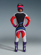 Motocross rider from behind wearing 2026 Techstar Knif Pants and matching jersey, light gray and black design with bright orange and purple accents, blue Alpinestars branding on the seat, purple boots and helmet, professional off-road racing gear set.
