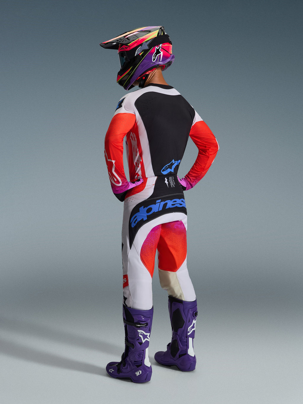 Motocross rider from the rear wearing the Alpinestars 2026 Techstar Knif Gear Set, white jersey with red sleeves and black panel, white pants with red and purple accents and blue Alpinestars branding, purple racing boots and matching helmet, professional off-road racing kit
