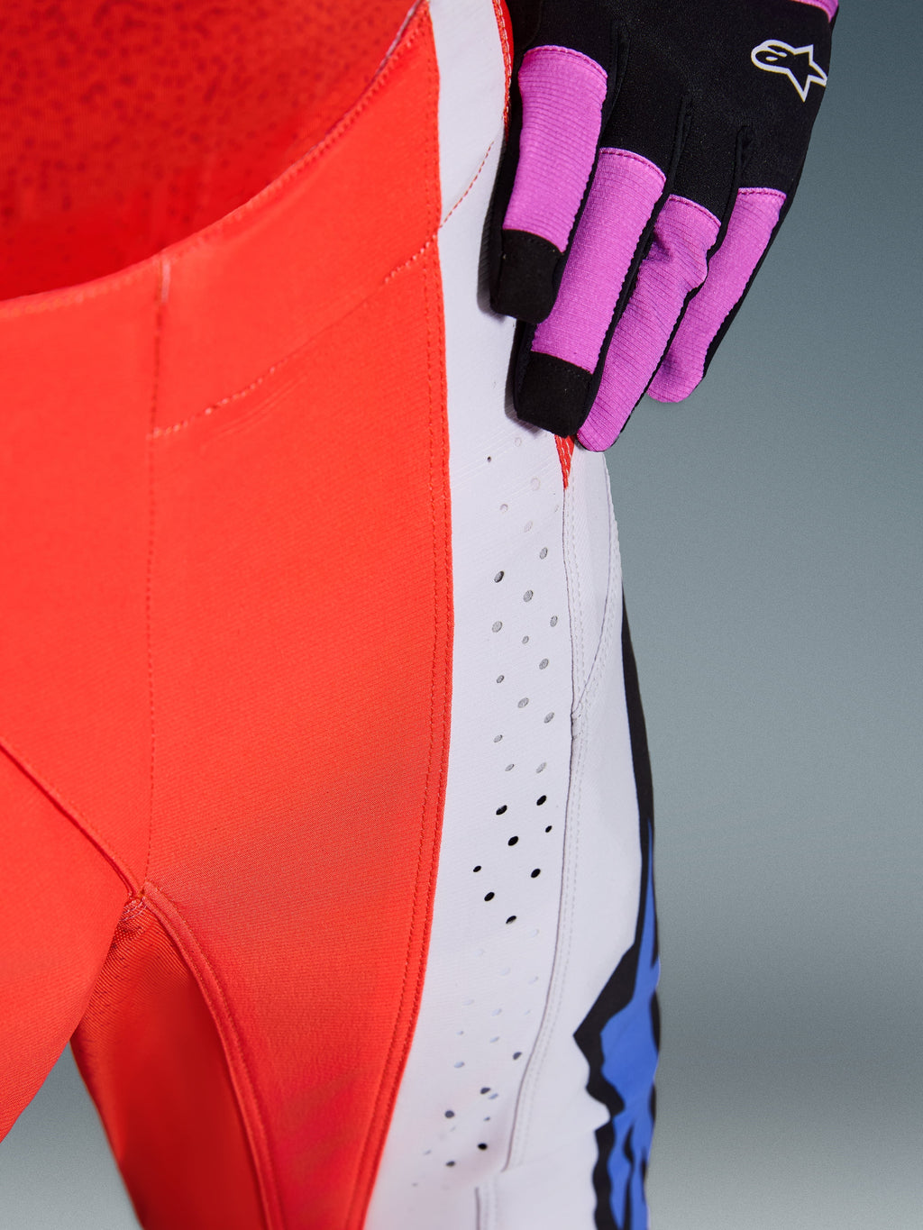 Alpinestars 2026 Techstar Knif Pants, Motocross Pants, Orange Fluo and Light Gray with blue accents, close-up showing laser-cut ventilation perforations on the thigh, worn with a black and purple motocross glove