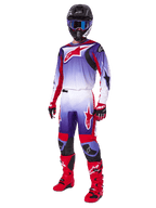 Pilote de motocross portant une tenue de course complète, pantalon Fluid Wurx 2025 et maillot assorti en violet, rouge vif et blanc, grand logo Alpinestars rouge, bottes et gants rouges, casque noir avec lunettes bleues, tenue coordonnée pour la course tout-terrain