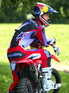 Pilote de motocross assis sur une moto tout-terrain rouge, portant un pantalon Alpinestars Fluid Wurx 2025 pourpre et rouge vif, maillot assorti, bottes Tech 10 blanches et noires, gants blancs et casque Red Bull, tenue de course tout-terrain complète en plein air