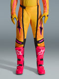 Pantalon Alpinestars Techstar Nomur 2026, pantalon de motocross, orange jaune et rose, porté avec des gants jaunes assortis et des bottes de course rose vif, avec empiècements noirs et logo Alpinestars rose audacieux, équipement technique de course tout-terrain