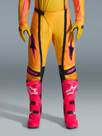 Pantalon Alpinestars Techstar Nomur 2026, pantalon de motocross, orange jaune et rose, porté avec des gants jaunes assortis et des bottes de course rose vif, avec empiècements noirs et logo Alpinestars rose audacieux, équipement technique de course tout-terrain