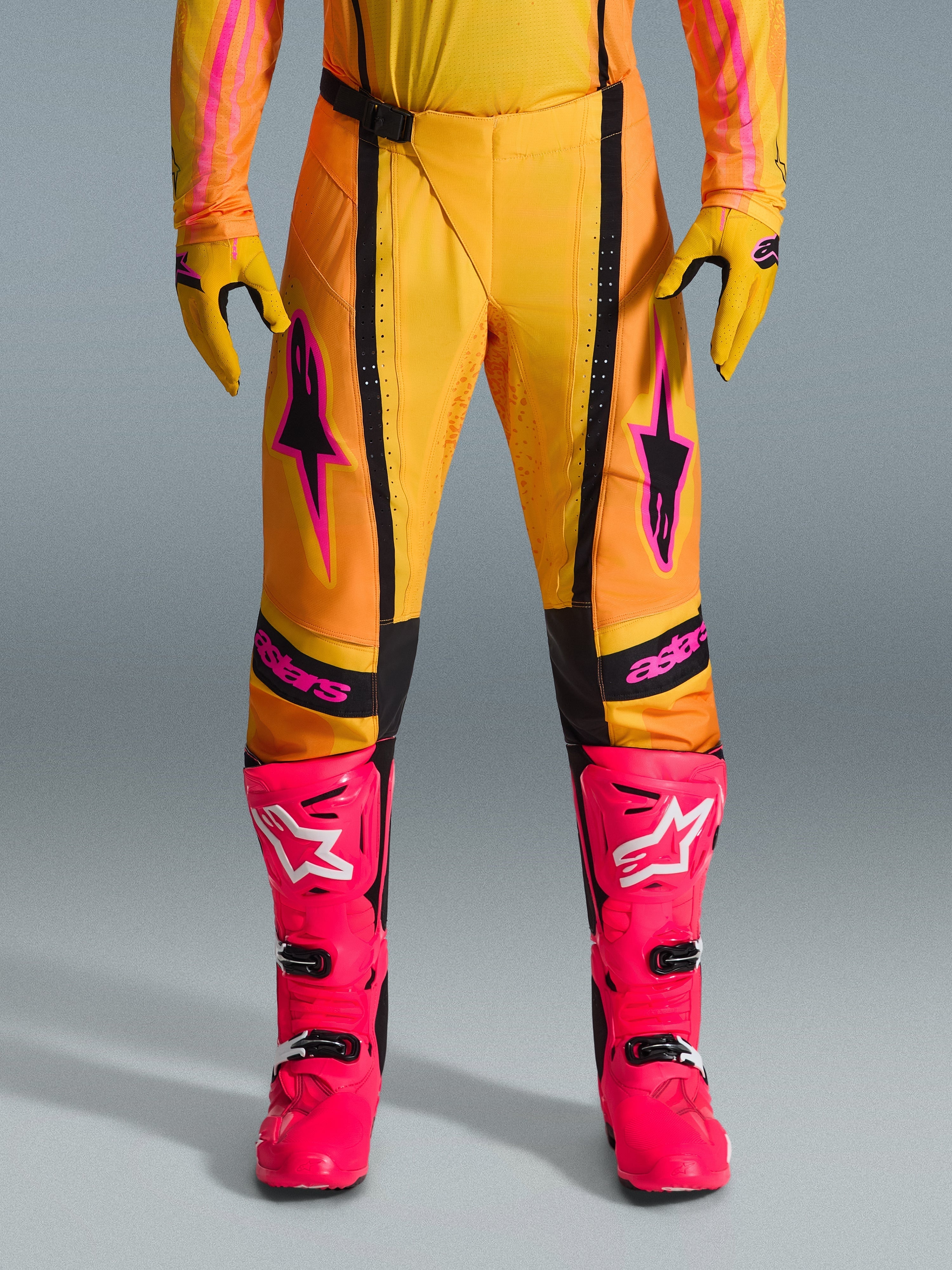 Pantalon Alpinestars Techstar Nomur 2026, pantalon de motocross, orange jaune et rose, porté avec des gants jaunes assortis et des bottes de course rose vif, avec empiècements noirs et logo Alpinestars rose audacieux, équipement technique de course tout-terrain