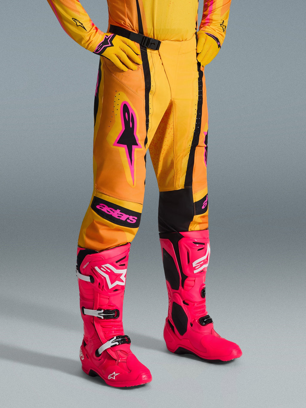 Pantalon Alpinestars Techstar Nomur 2026, pantalon de motocross, orange, jaune et rose, porté avec un maillot et des gants jaunes assortis et des bottes rose vif, coupe de course athlétique avec logos roses et boucle magnétique noire, équipement éclatant pour la compétition tout-terrain