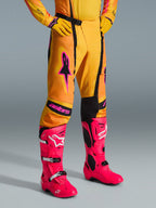 Pantalon Alpinestars Techstar Nomur 2026, pantalon de motocross, orange, jaune et rose, porté avec un maillot et des gants jaunes assortis et des bottes rose vif, coupe de course athlétique avec logos roses et boucle magnétique noire, équipement éclatant pour la compétition tout-terrain