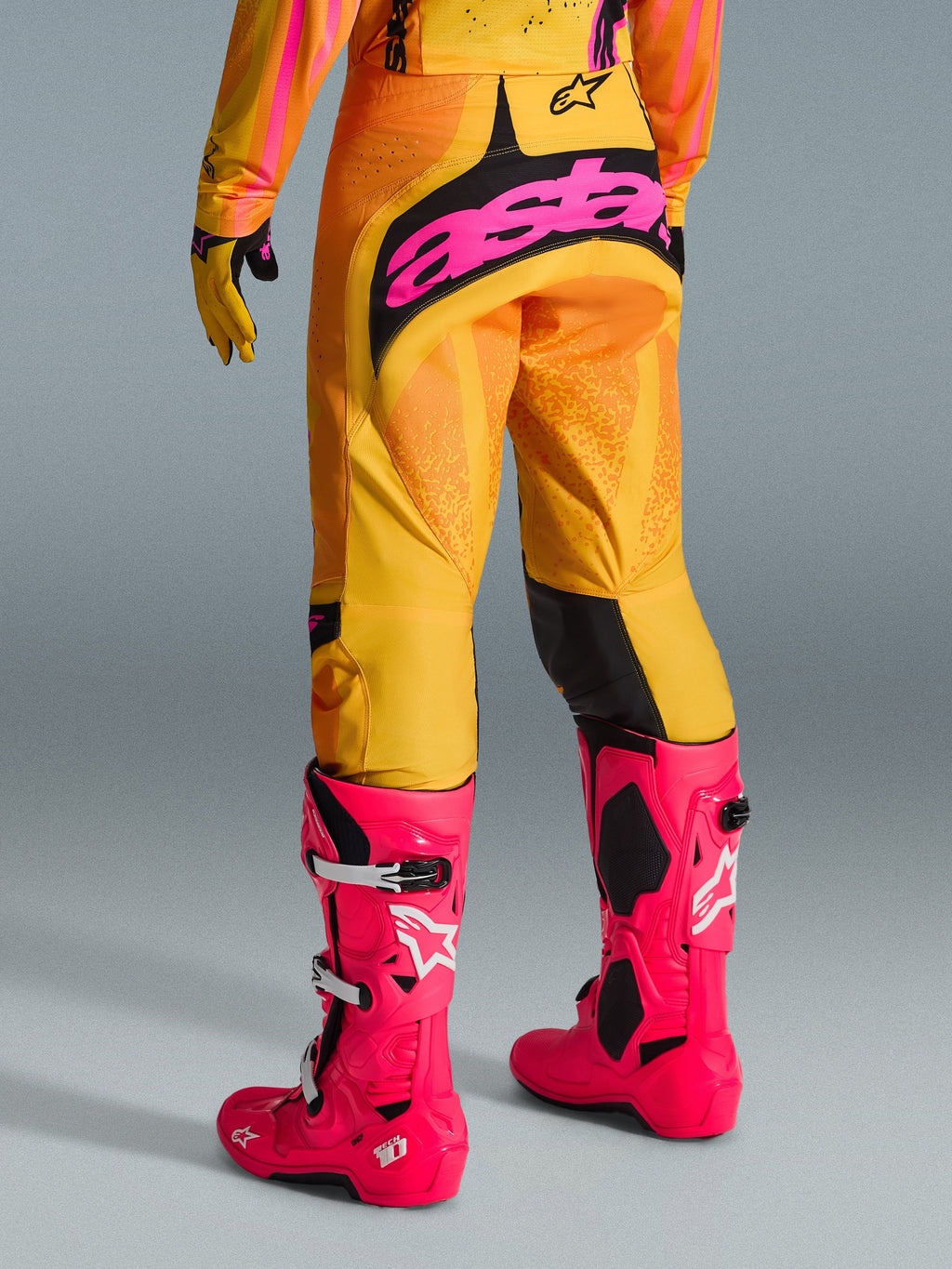 Pantalon Alpinestars Techstar Nomur 2026, pantalon de motocross, orange, jaune et rose, vue arrière, arborant un grand logo 'astars' rose sur l'assise, associé à des bottes Tech 10 rose vif et un maillot assorti, équipement de course haute performance pour motocross