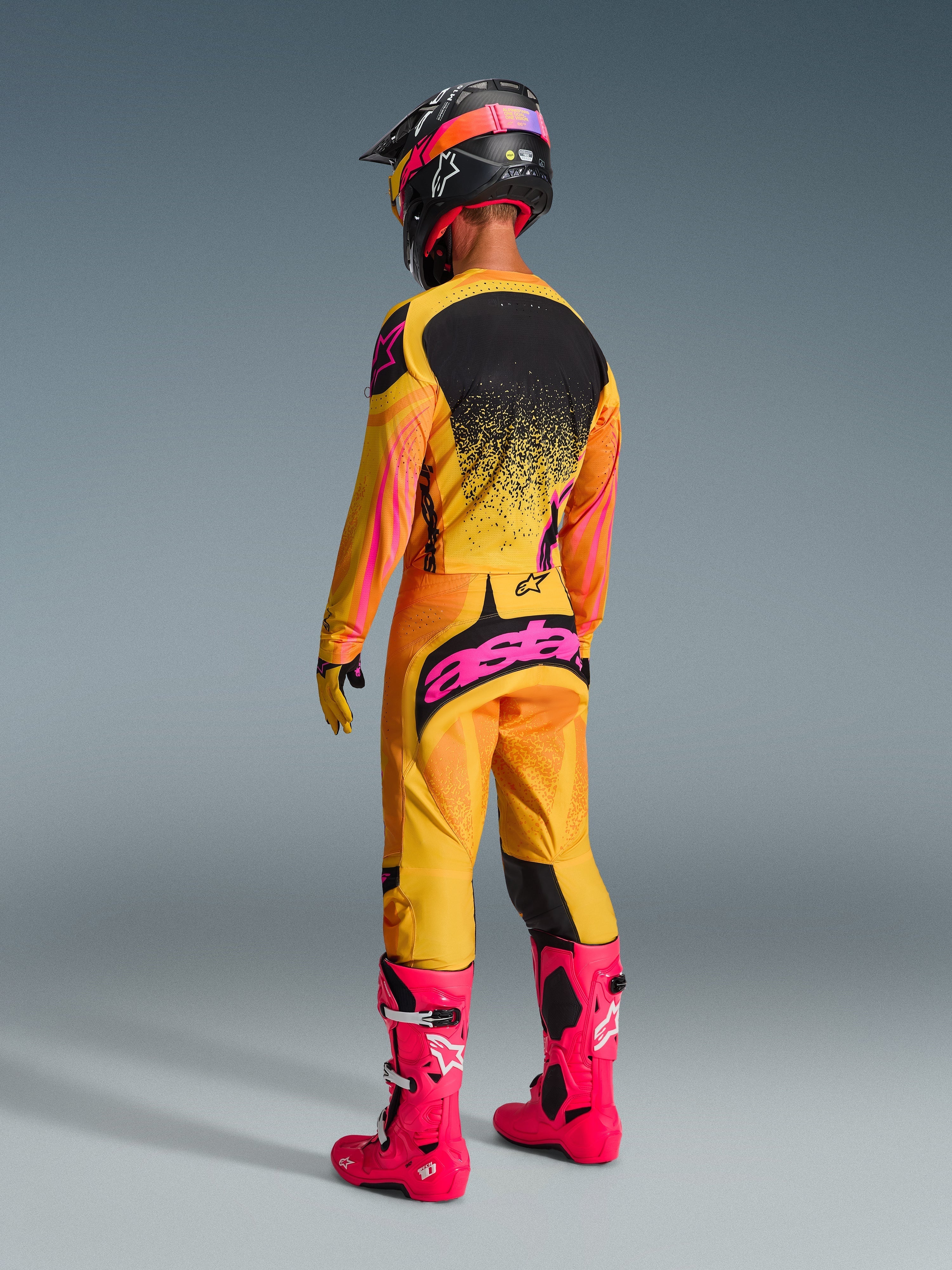 Pilote de motocross vu de dos, portant un équipement de course complet, pantalon Techstar Nomur 2026 en orange, jaune et rose, marquage 'astars' rose vif sur l'assise, maillot assorti, bottes rose vif et casque noir avec accents roses, tenue de compétition tout-terrain coordonnée.