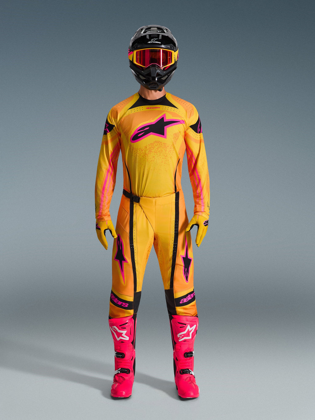 Pilote de motocross portant une tenue complète Alpinestars Techstar Nomur 2026, maillot et pantalon orange, jaune et rose, gants assortis jaune et rose, bottes de course rose vif, et casque noir avec lunettes roses, ensemble de motocross haute performance coordonné.