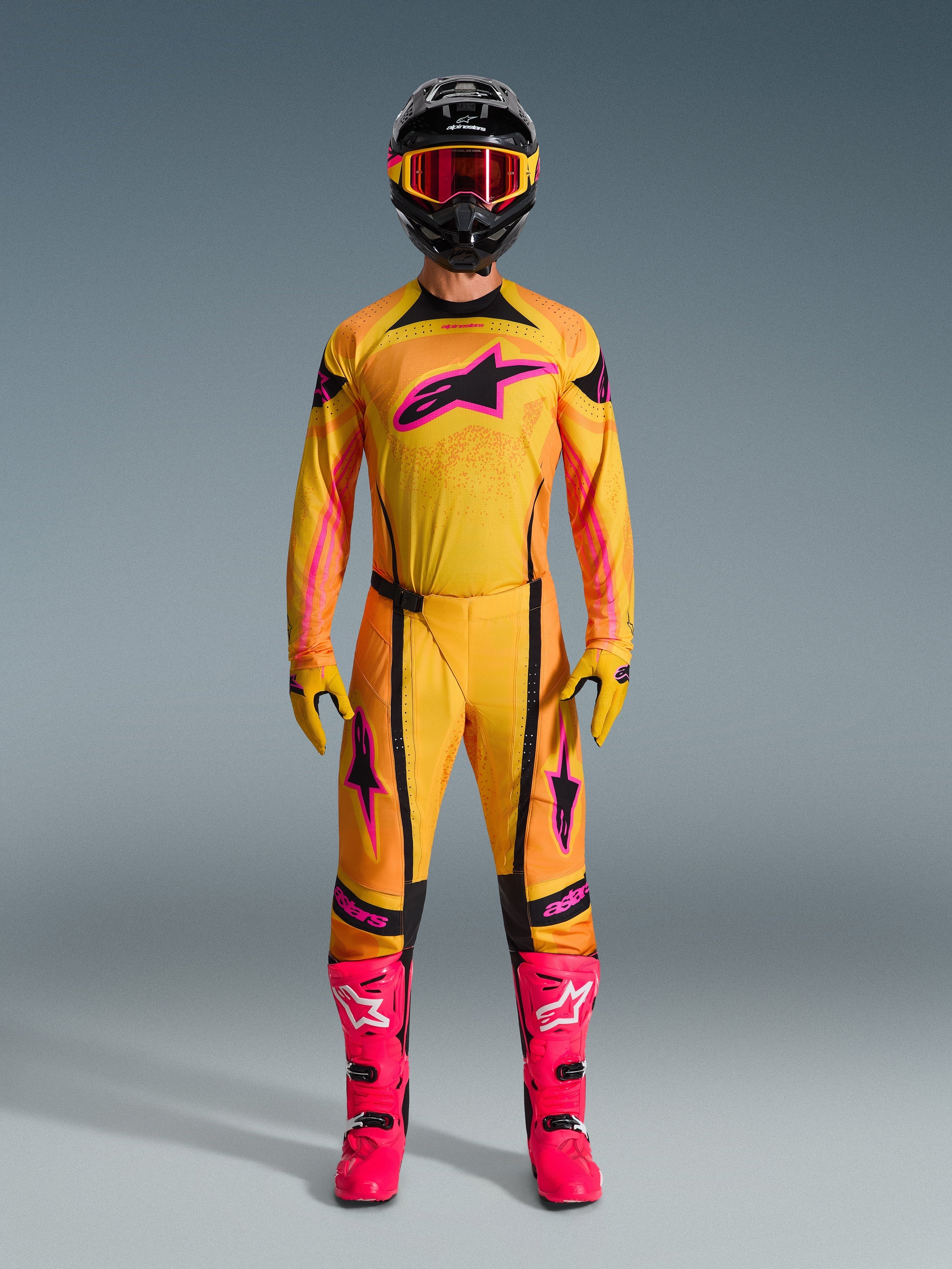 Pilote de motocross portant une tenue complète Alpinestars Techstar Nomur 2026, maillot et pantalon orange, jaune et rose, gants assortis jaune et rose, bottes de course rose vif, et casque noir avec lunettes roses, ensemble de motocross haute performance coordonné.