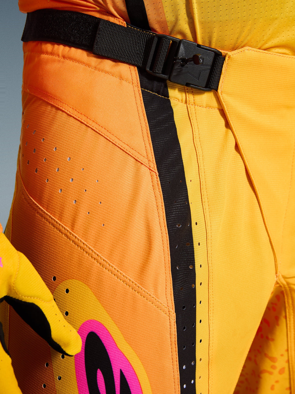 Alpinestars 2026 Techstar Nomur Pants, pantalon de motocross, orange, jaune et rose, gros plan sur la zone de la taille présentant la fermeture par boucle magnétique SX2, sangle de réglage noire et perforations de ventilation découpées au laser sur un matériau ripstop durable