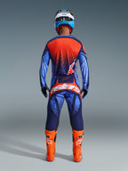 Motocross rider seen from the back wearing Alpinestars 2026 Techstar Nomur Pants in Dark Navy, Orange, and UCLA Blue, styled with a matching jersey and bright orange boots, professional off-road racing gear set.