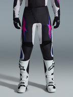 Alpinestars 2026 Fluid Grid Pants, pantalon de motocross, noir avec motifs d'étoiles violets et bleus, porté avec des bottes de motocross blanches et des gants noirs, avec empiècements renforcés et coupe sportive tout-terrain, fond gris