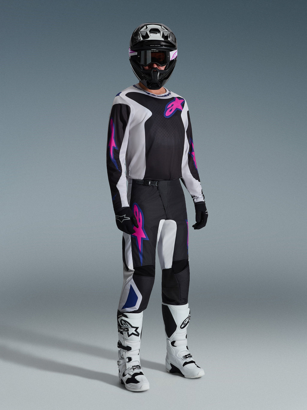 Pilote de motocross portant une tenue de course complète, pantalon Alpinestars 2026 Fluid Grid en noir, gris clair et violet, maillot assorti avec graphismes roses et bleus, casque et lunettes noirs, bottes blanches et noires, et gants noirs, équipement coordonné pour la course tout-terrain