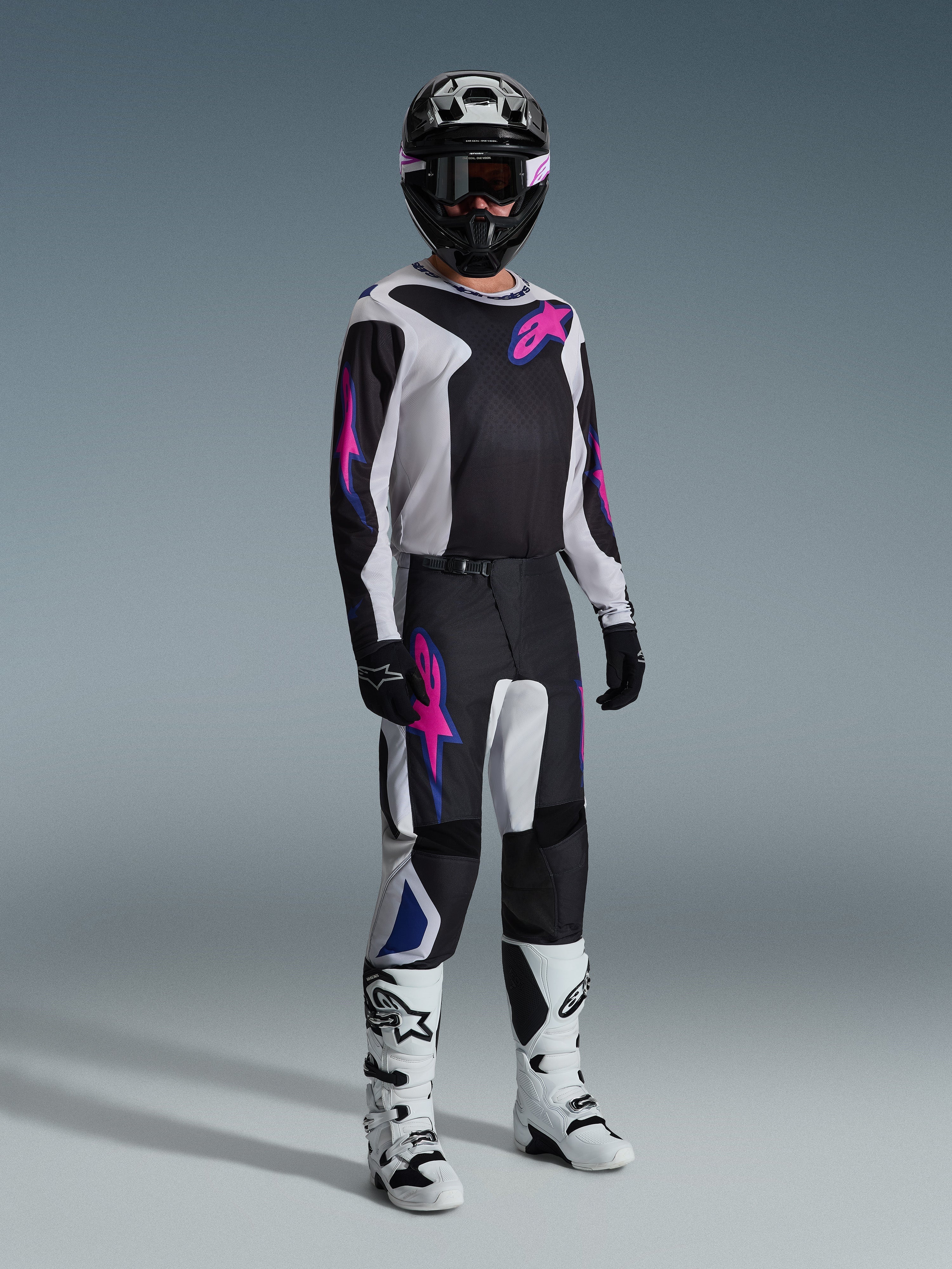 Pilote de motocross portant une tenue de course complète, pantalon Alpinestars 2026 Fluid Grid en noir, gris clair et violet, maillot assorti avec graphismes roses et bleus, casque et lunettes noirs, bottes blanches et noires, et gants noirs, équipement coordonné pour la course tout-terrain