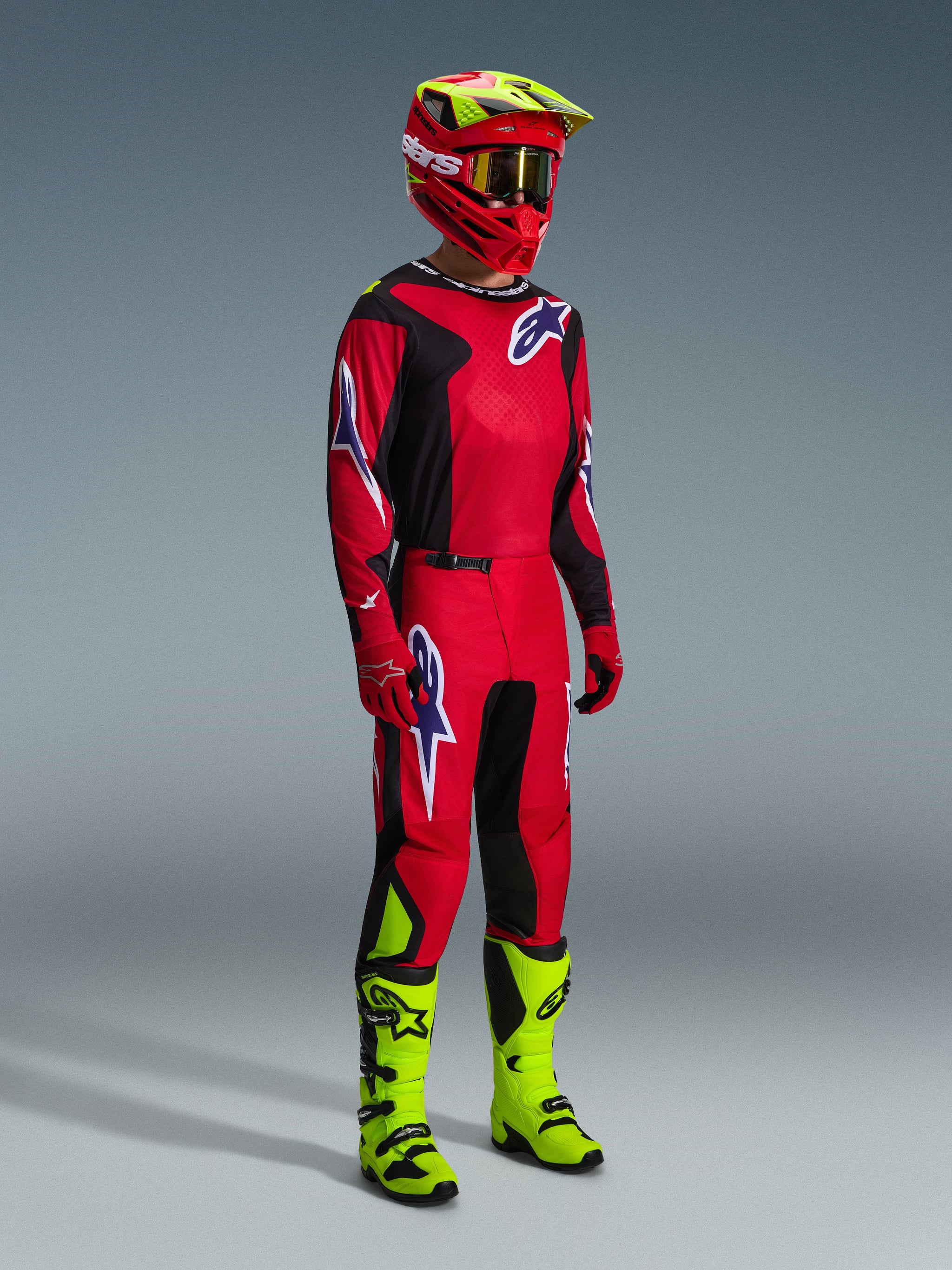 Motocross rider wearing the 2026 Fluid Grid Pants in Red Black Purple, complete racing gear with matching jersey, bright red and fluo yellow helmet, and fluo yellow boots, coordinated off-road racing outfit featuring bold Alpinestars branding