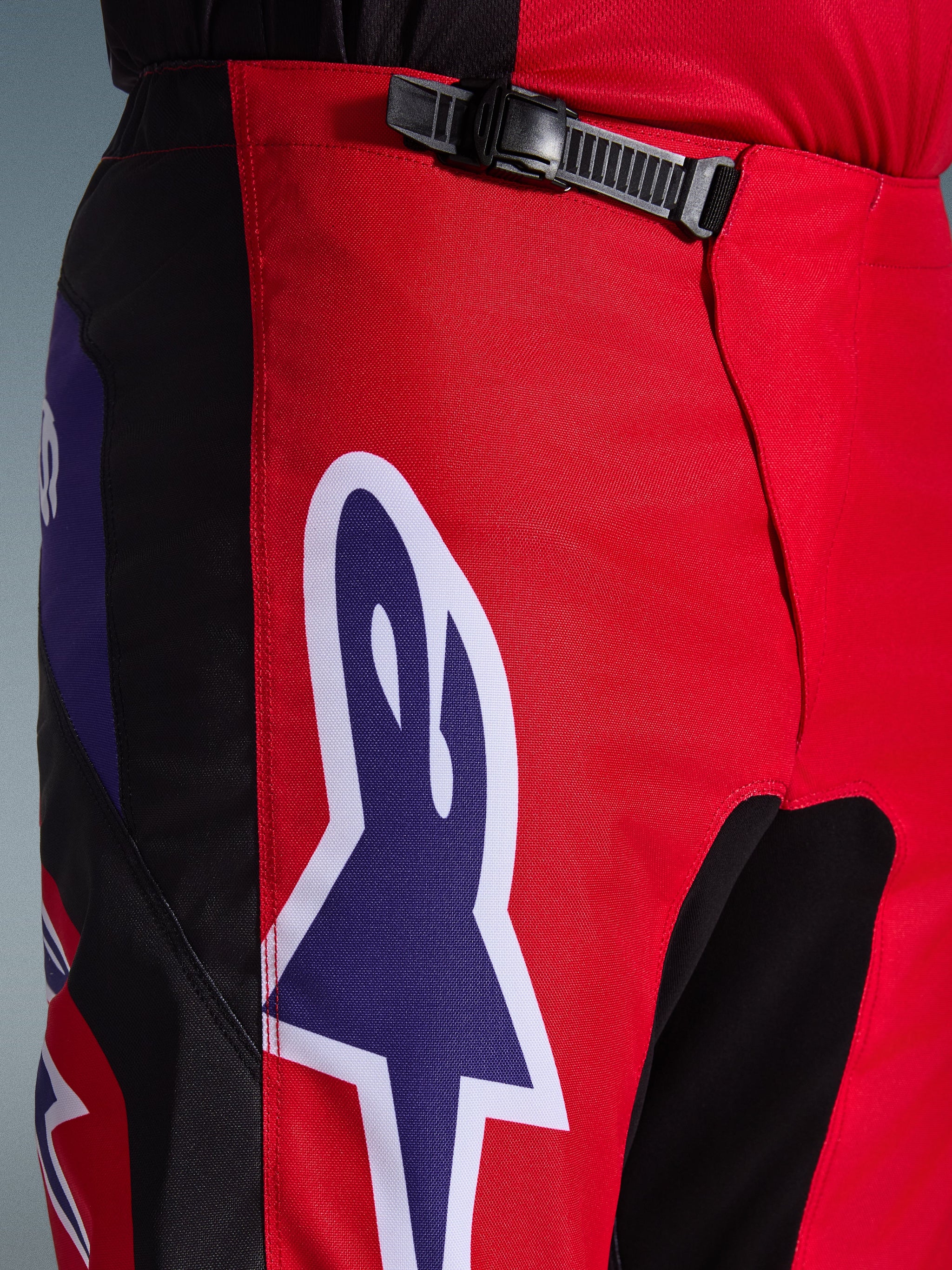 Alpinestars 2026 Fluid Grid Pants, Motocross Pants, Red Black and Purple, close-up of the waist and hip area featuring a black ratchet closure system and a large purple Alpinestars star logo with white outline, durable textured fabric for off-road racing.