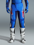 Pantalon Alpinestars 2026 Fluid Grid, pantalon de motocross, bleu et rouge, avec des logos en forme d'étoile rouges et blancs sur les cuisses, porté avec des gants bleus assortis et des bottes tout-terrain blanches, coupe athlétique avec des panneaux durables pour la course.
