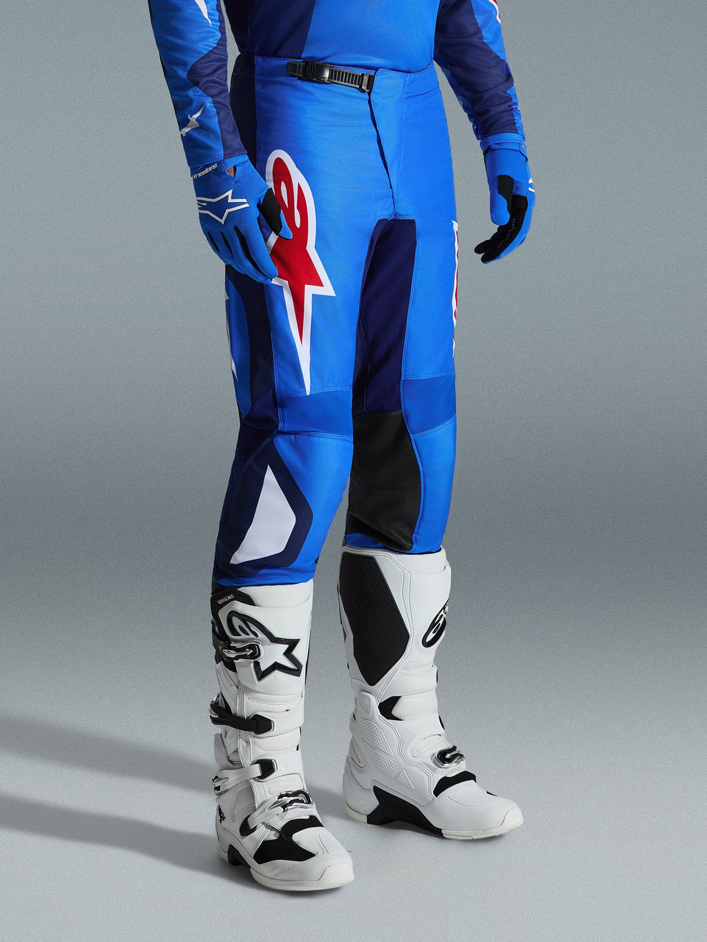 Alpinestars 2026 Fluid Grid Pants, pantalon de motocross, bleu et rouge, porté avec des bottes de motocross blanches et des gants bleus, arborant un grand logo étoile rouge sur la cuisse et des panneaux intérieurs noirs aux genoux, conçu pour la conduite tout-terrain