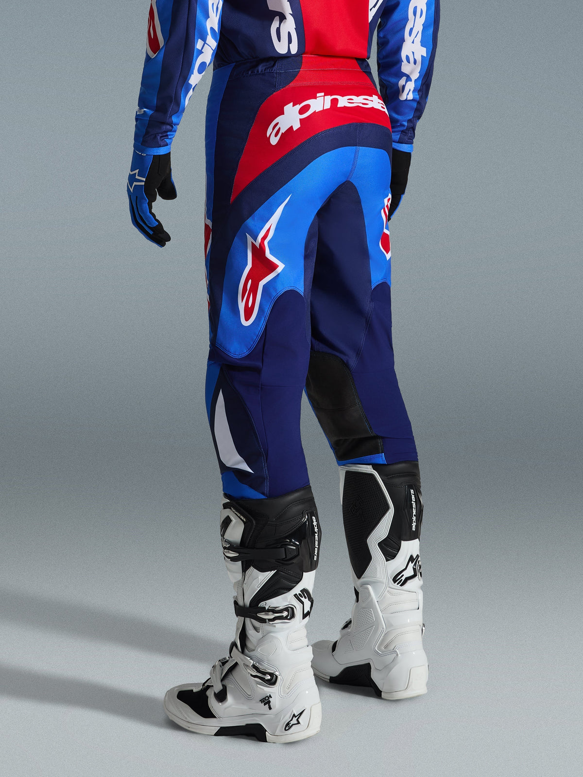 Pantalon Alpinestars Fluid Grid 2026, pantalon de motocross, bleu et rouge, vue arrière montrant le marquage Alpinestars audacieux et le design ergonomique, porté avec des bottes Tech 7 blanches et noires et un maillot assorti, équipement haute performance pour la conduite tout-terrain
