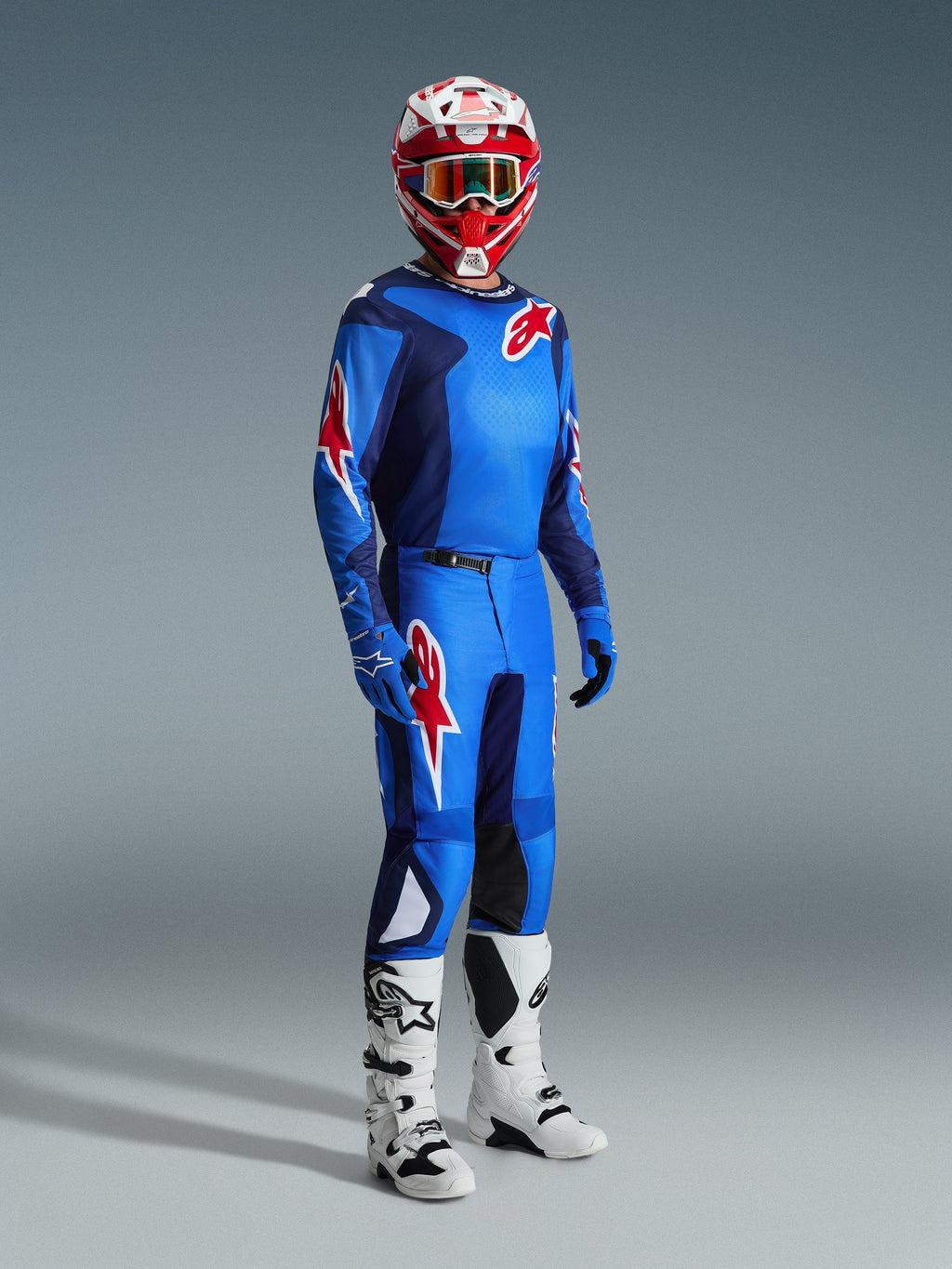 Pilote de motocross portant un équipement de course complet Alpinestars, maillot bleu et rouge avec motif quadrillé et logo, pantalon Fluid Grid 2026 bleu et rouge avec empiècements intérieurs noirs aux genoux, casque rouge et blanc avec lunettes, bottes blanches et noires, gants bleus, tenue coordonnée pour la course tout-terrain