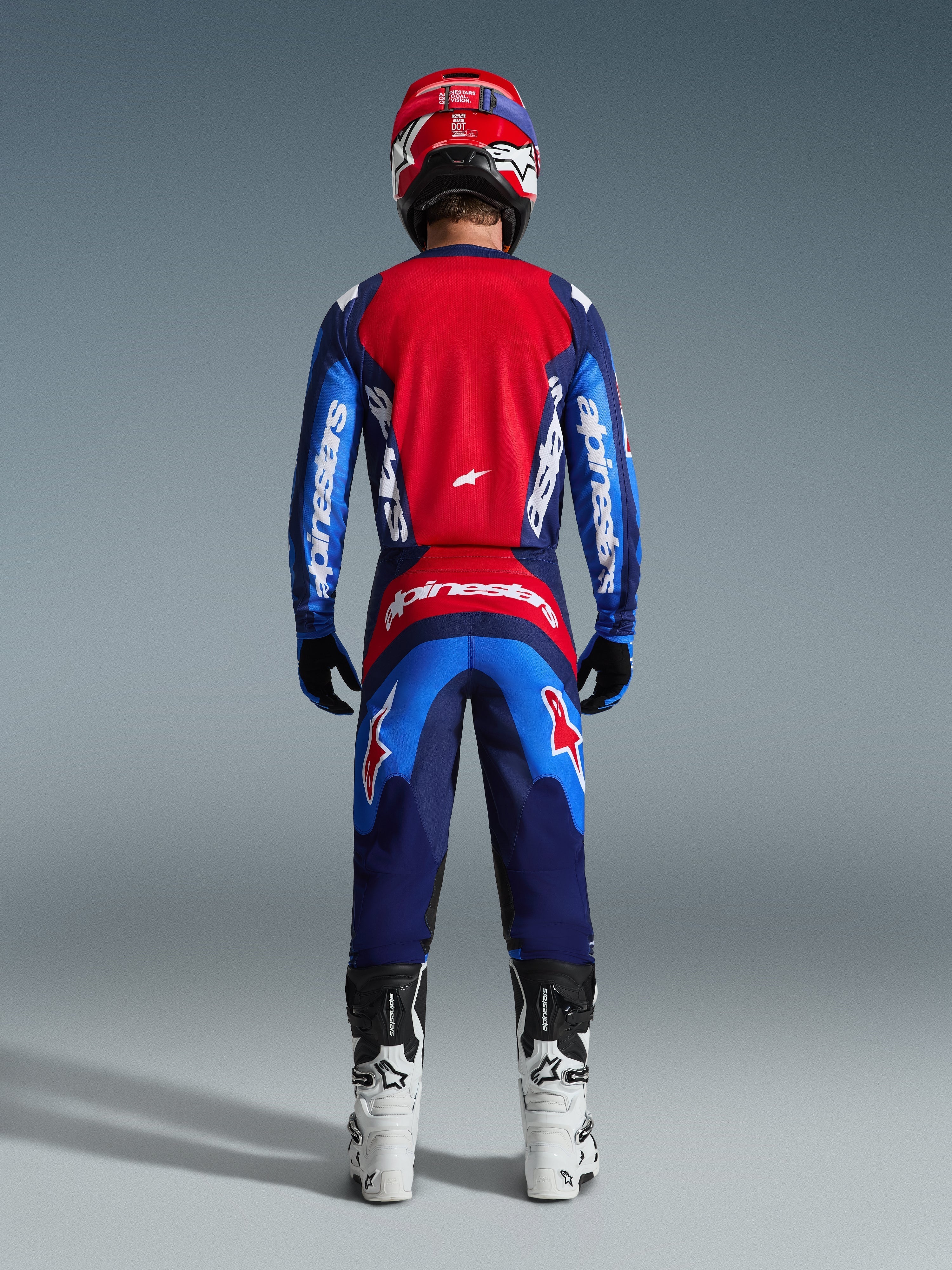 Pilote de motocross vu de dos portant un équipement de course complet, pantalon Fluid Grid 2026 en bleu et rouge, maillot assorti avec logo Alpinestars, casque rouge et bleu, bottes blanches et noires, tenue coordonnée pour le tout-terrain