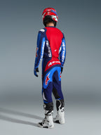 Pilote de motocross vu de dos portant un pantalon Alpinestars Fluid Grid 2026 Bleu Rouge, maillot bleu et rouge assorti avec logos blancs, casque rouge et blanc, bottes blanches et noires, tenue coordonnée pour la course tout-terrain