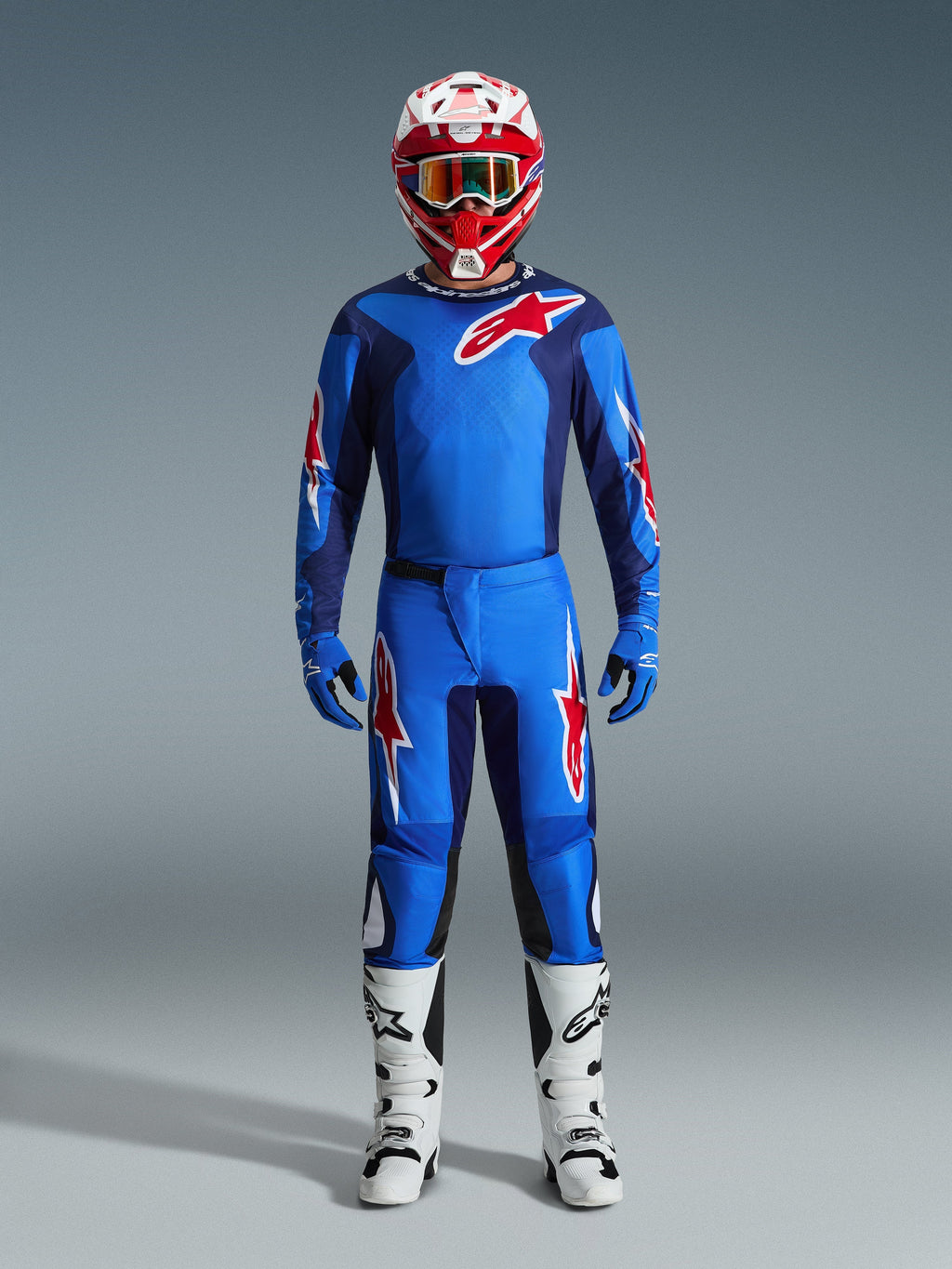 Pilote de motocross portant le pantalon Alpinestars Fluid Grid 2026 bleu et rouge, maillot bleu assorti avec un logo Astars rouge, casque rouge et blanc avec lunettes, gants bleus et bottes de motocross blanches, équipement de course tout-terrain complet et coordonné.