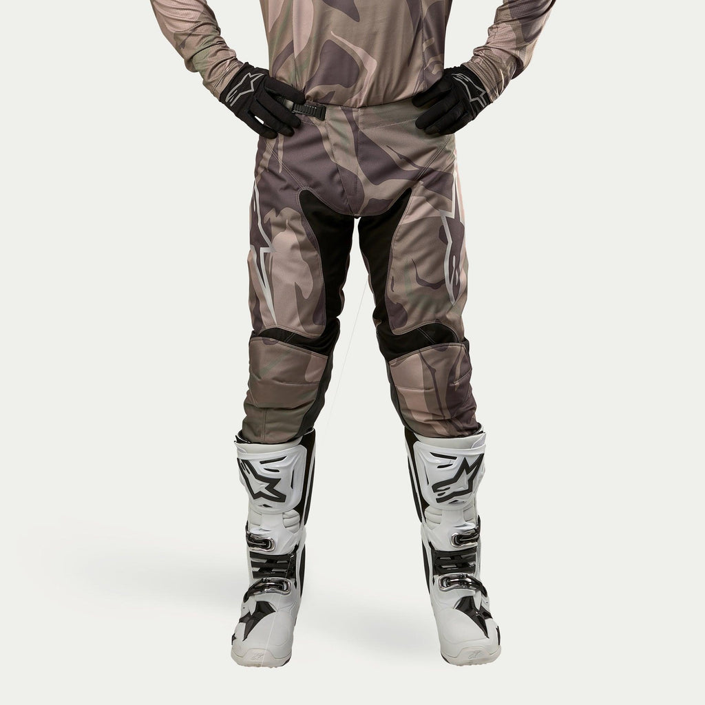 Alpinestars 2024 Racer Tactical Pants, pantalon de motocross, camouflage vert militaire et marron, avec de grands logos Alpinestars blancs sur les cuisses et des empiècements noirs à l'intérieur des jambes, porté avec des bottes de motocross blanches et noires et des gants noirs, maillot de camouflage assorti, vue de face.