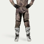 Alpinestars 2024 Racer Tactical Pants, pantalon de motocross, camouflage vert militaire et marron, avec de grands logos Alpinestars blancs sur les cuisses et des empiècements noirs à l'intérieur des jambes, porté avec des bottes de motocross blanches et noires et des gants noirs, maillot de camouflage assorti, vue de face.